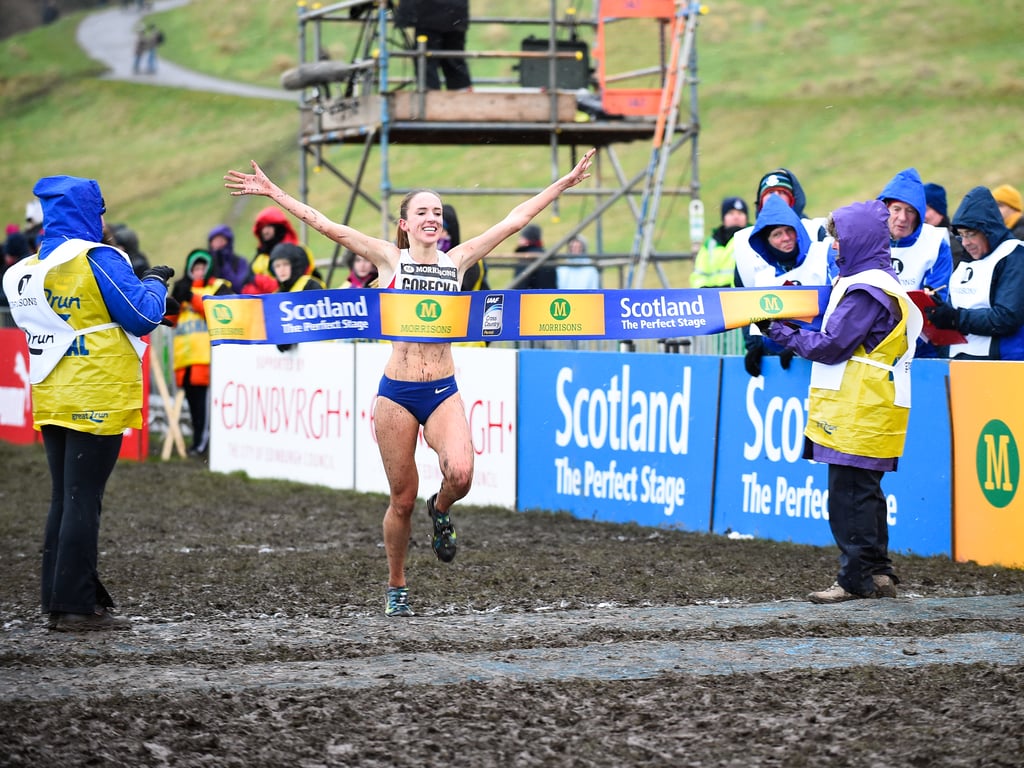 A woman crossing the finish line of a 6K cross-country running race with her hands up in the air.